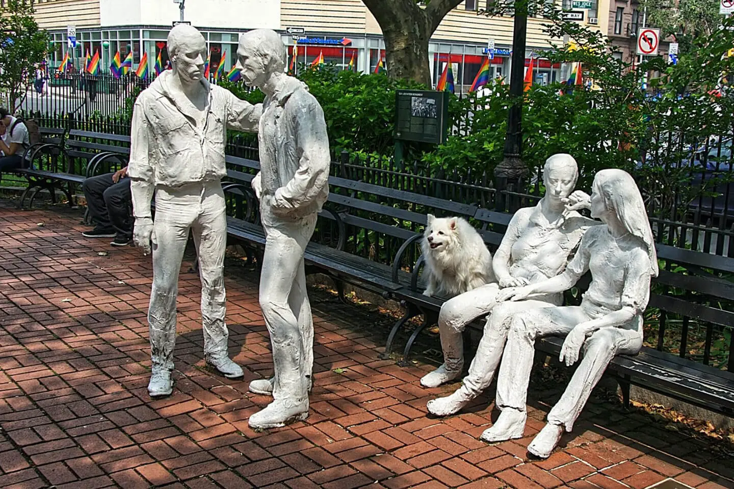 Life-sized statues depicting a group of figures in mid-conversation with a dog, situated in a public space with benches and trees in Manhattan.
