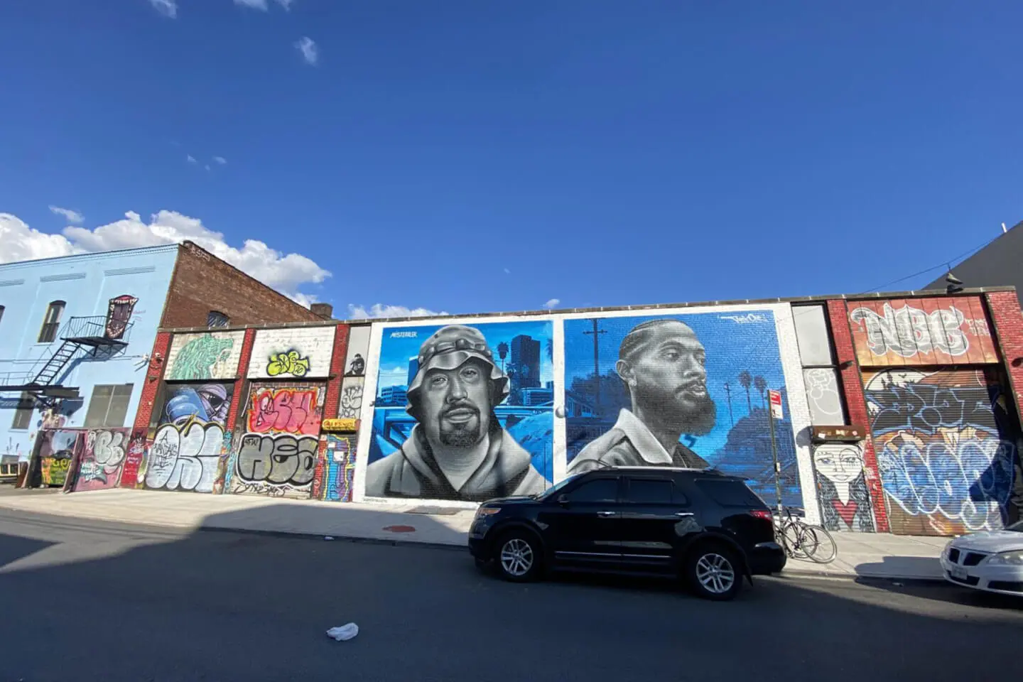 A colorful street mural of two men's faces on an urban building wall in Manhattan with parked cars in front.