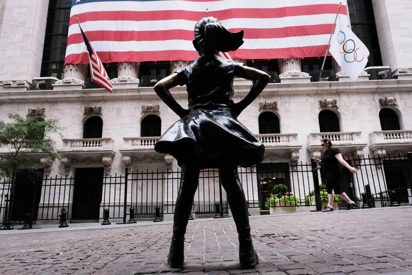 A statue of a girl with hands on hips standing in front of the New York Stock Exchange in Manhattan, under a large American flag.