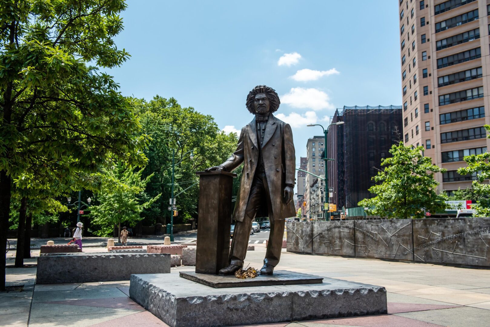 Statue of frederick douglass in a city park with trees and buildings in the background.