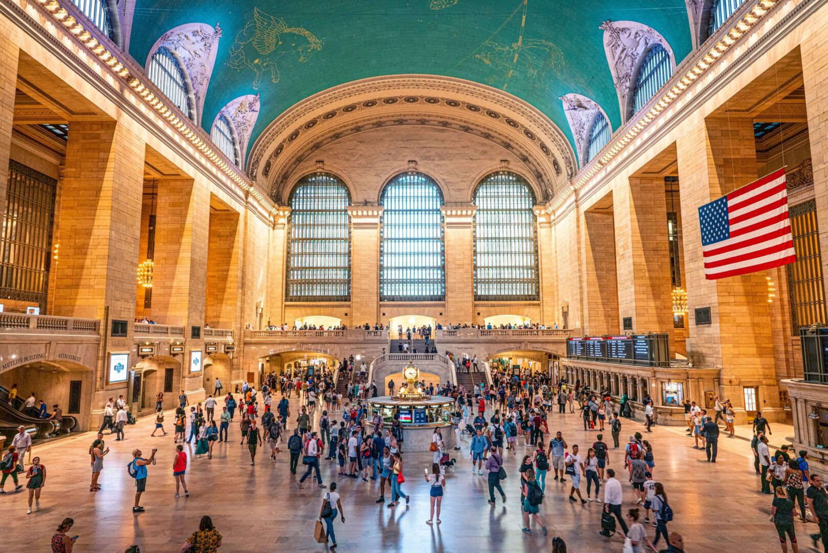 Grand central terminal bustling with activity beneath an american flag and the ornate celestial ceiling.