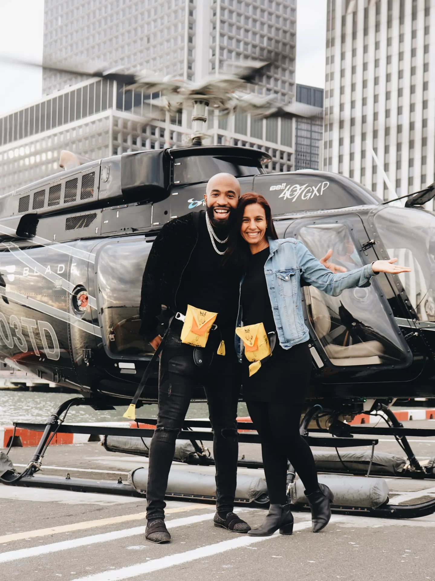 A smiling man and woman standing in front of a luxury helicopter with the cityscape in the background.