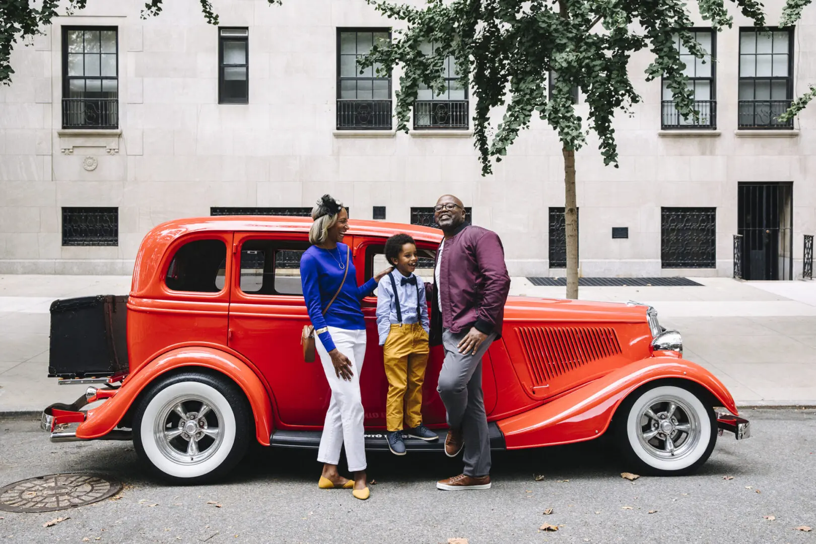 A family dressed in luxury, stylish attire standing next to a classic red car on a city street.